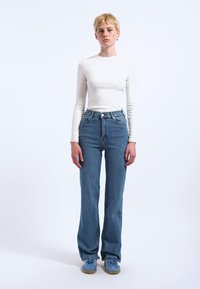 Hoch geschnittene blaue Jeans mit geradem Bein, aus leichtem Denim-Material. Kombiniert mit einem taillierten weißen Langarmshirt und blauen Turnschuhen.