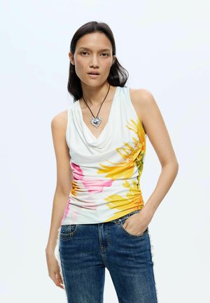 Femme portant un débardeur blanc avec de grandes fleurs jaunes et roses, un jean bleu, et un collier pendentif cœur en argent.