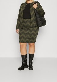 Giacca verde oliva con motivo a onde, abbinata a un top nero e una gonna coordinata. Stivali neri al ginocchio e una borsa nera trapuntata.