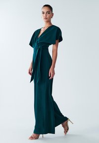 Teal wrap jumpsuit with short flutter sleeves, a tie waist, and a straight leg design; paired with gold strappy high-heeled sandals.