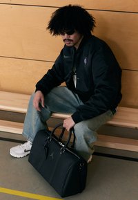 Sort læder duffeltaske med sølvbeslag, med logo og mærkat. Model iført sort jakke og solbriller, siddende på en træbænk.