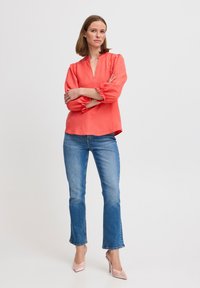 Koralfarvet bluse med lange ærmer og puffede ærmer, notch-udskæring og struktureret stof, bæres med blå jeans og lyserøde højhælede sko.