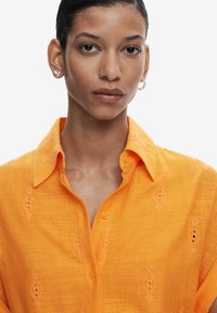 Camicia arancione ricamata con colletto, caratterizzata da motivi decorativi e chiusura con bottoni, realizzata in tessuto leggero.