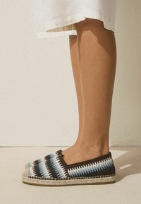 Gambe con vestito bianco e espadrillas slip-on con motivo a zigzag blu, bianco e nero, in piedi su pavimento chiaro contro muro bianco.