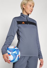 Sivi sportski zip-up top s crnim i narančastim detaljima, dugim rukavima i glatkom teksturom. Drži plavi i bijeli lopta sa uzorkom.