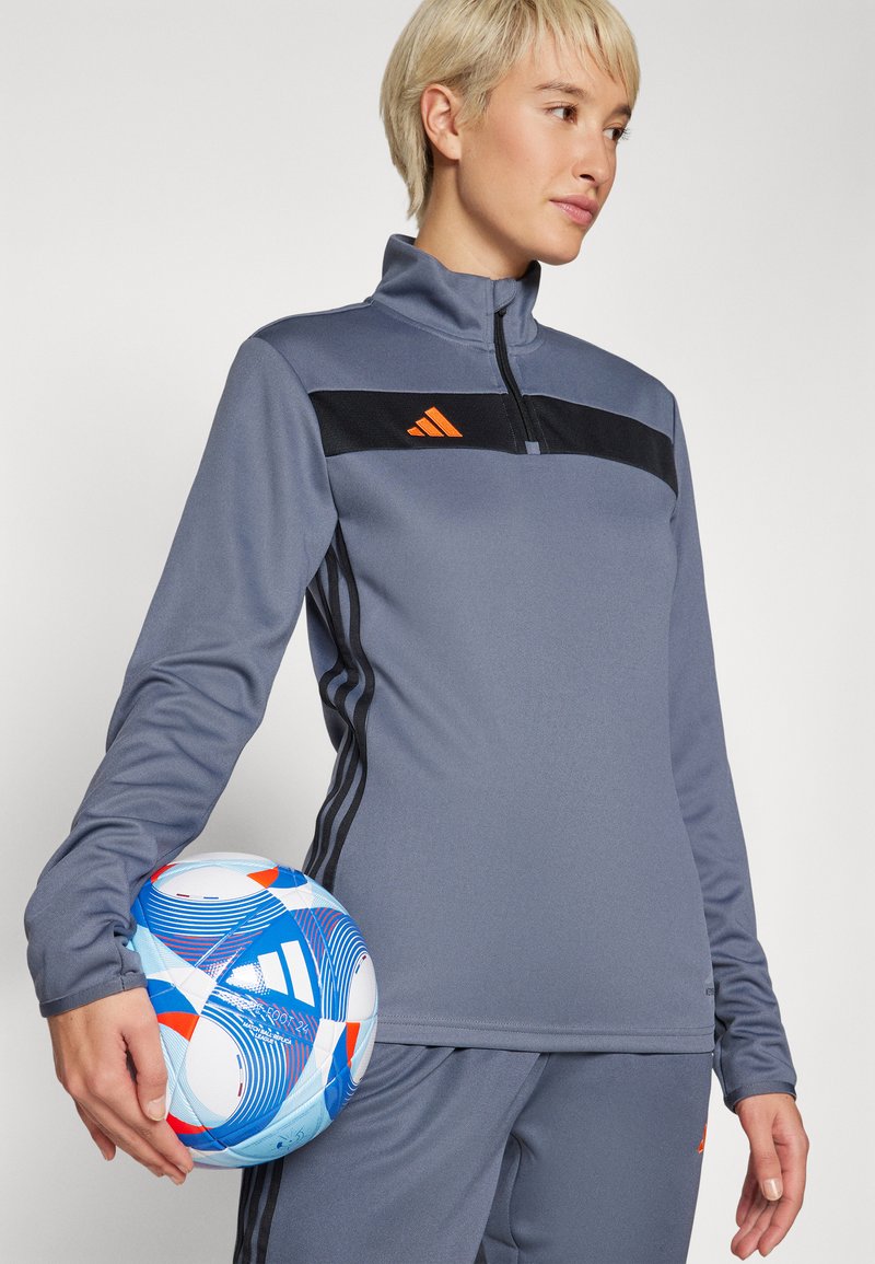 Grauer sportlicher Reißverschluss-Oberteil mit schwarzen und orangefarbenen Akzenten, langen Ärmeln und glatter Textur. Hält einen blau-weiß gemusterten Fußball.