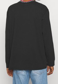 Schwarzes langärmeliges T-Shirt aus weichem Stoff, mit lockerer Passform, Rundhalsausschnitt und rot-weißen Mustern am Kragen.