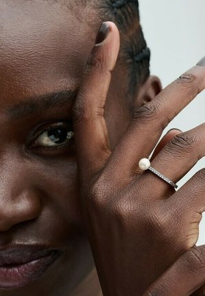 Perlring an einer Hand; Silberband mit zartem Design und einer runden weißen Perle; der Hautton ist reich und tief strukturiert.