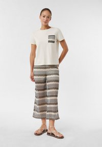 Crème shirt met korte mouwen en een zwarte zigzagzak, gecombineerd met een patroon van tan en zwarte wijde broek. Het model draagt tan sandalen met banden.