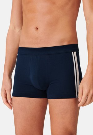 Boxer en bleu marine fabriqué dans un tissu doux, présentant une coupe ajustée et deux bandes blanches horizontales sur les côtés. Taille élastique.