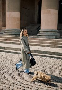 Kobieta w płaszczu przeciwdeszczowym spacerująca małym, puszystym psem na smyczy po brukowanej ulicy przed kamiennymi schodami i dużymi kolumnami.