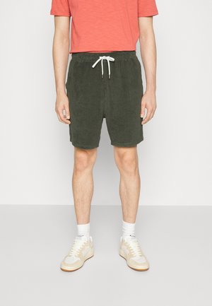 Männliches Model trägt dunkelgrüne Shorts mit Kordelzug, korallenfarbenes Kurzarmhemd, weiße Sportsocken und beige Sneakers, steht vor einem einfarbigen Hintergrund.
