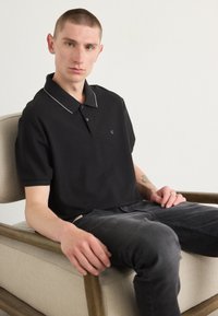 Svart pikétröja med grå krage och korta ärmar, med logotyp på bröstet. Bärs med mörka jeans medan personen sitter på en ljus stol.