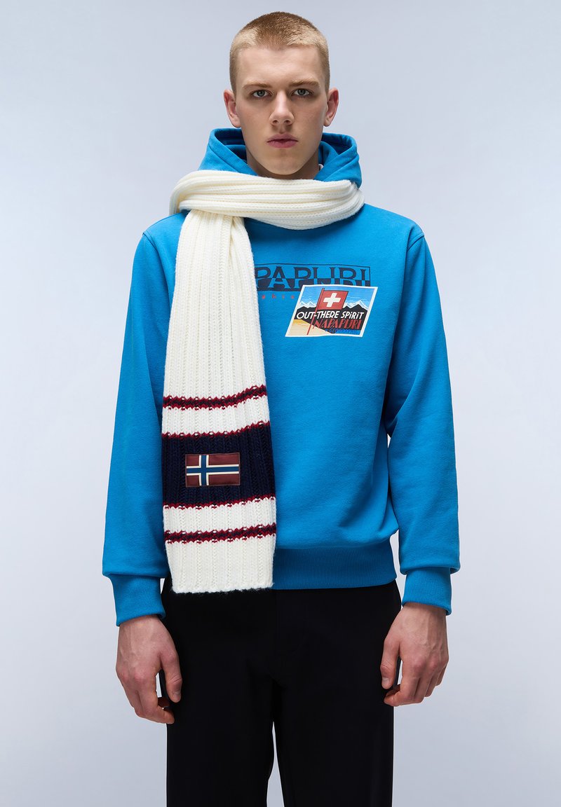 Blå hættetrøje med logo i bomuldsstof; cremefarvet strikket tørklæde med marineblå og røde striber, med et norsk flag patch; sorte bukser.