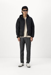 Schwarze gepolsterte Jacke mit Kapuze, darunter ein cremefarbenes Hemd, graue, schmal zulaufende Hose und schwarze Sonnenbrille, kombiniert mit weißen Sneakers mit schwarzen Streifen.