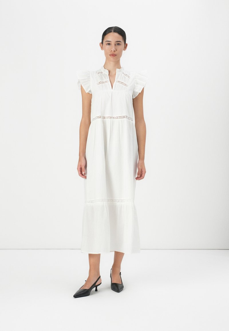 Rochie albă din bumbac cu mâneci volănoi, detalii din dantelă la talie, tiv în straturi și guler înalt, asortată cu pantofi cu toc slingback negri.