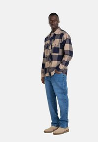 Camisa de franela a cuadros en beige y azul marino con dos bolsillos en el pecho, combinada con jeans de color azul claro y botas color marrón claro, de pie contra un fondo blanco.