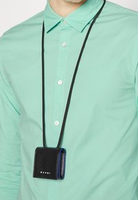 Borsetta in pelle nera con dettagli blu, indossata su una tracolla nera, esposta su una maglietta verde menta. Presenta il nome del marchio "MARNI" in bianco.