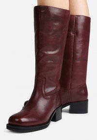 Botas de couro bordeaux até ao joelho com um acabamento suave, ponta quadrada e salto bloco. Apresentam costura decorativa ao longo dos lados.