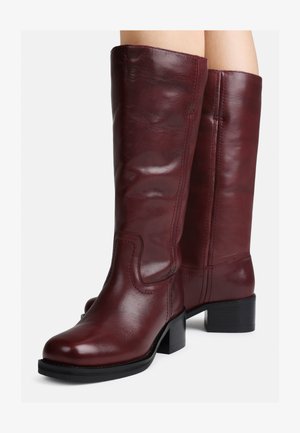 Botas de couro bordeaux até ao joelho com um acabamento suave, ponta quadrada e salto bloco. Apresentam costura decorativa ao longo dos lados.