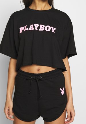 Femme portant un t-shirt noir court avec l'inscription "PLAYBOY" en rose et un short noir assorti avec le logo du lapin Playboy rose sur le côté gauche.