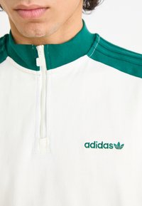 Λευκή ζακέτα με φερμουάρ, με πράσινη περιλαβή και ρίγες στους ώμους. Διαθέτει κεντημένο λογότυπο "adidas" σε πράσινο χρώμα στο στήθος. Μαλακή υφή υφάσματος.