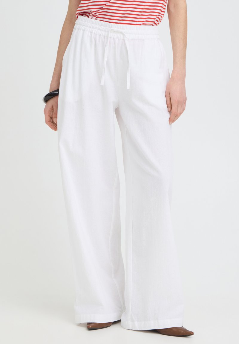 Personne portant un pantalon blanc ample avec cordon, un haut rayé rouge et blanc, des chaussures marron à bouts pointus et un bracelet noir au poignet gauche.
