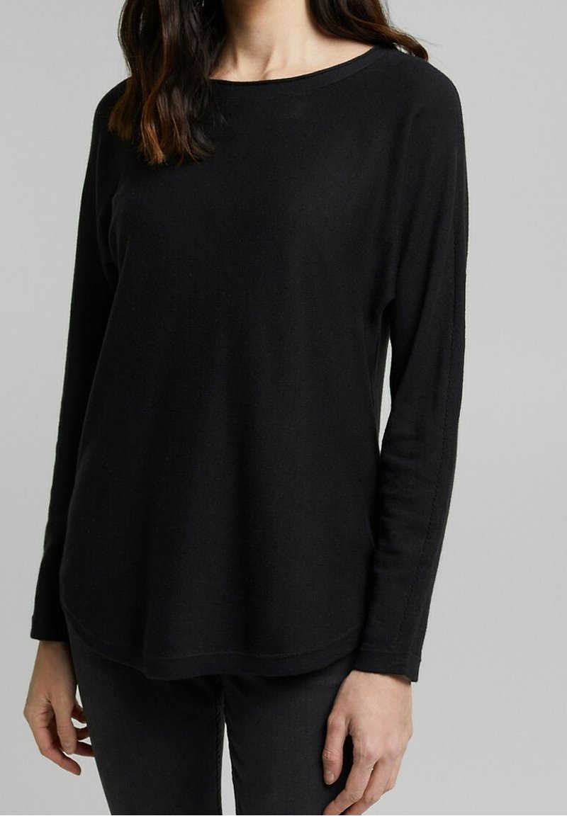 Femme portant un chemisier noir à manches longues et un pantalon noir, debout contre un fond gris, les mains détendues le long du corps.