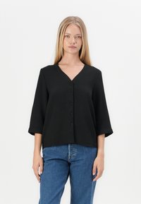 Svart knappebluse med V-hals og trekvartlange ermer, laget av teksturert stoff. Kombinert med blå denimjeans.