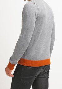 Grau gestrickter Pullover mit rundem Halsausschnitt, geripptem orangenem Saum und Bündchen, kombiniert mit dunkelgrauen Jeans. Weiche Textur, taillierte Passform.