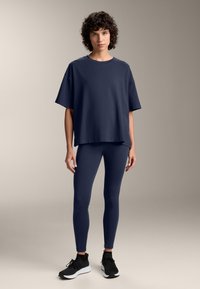 Tricou oversized bleumarin asortat cu colanți bleumarin și pantofi sport negri. Material neted, ușor, cu un fit relaxat.