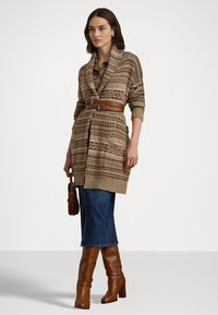 Cardigã de malha em tons terrosos com padrões geométricos, cinto na cintura, combinado com uma saia de ganga e botas de salto alto castanhas até ao joelho.