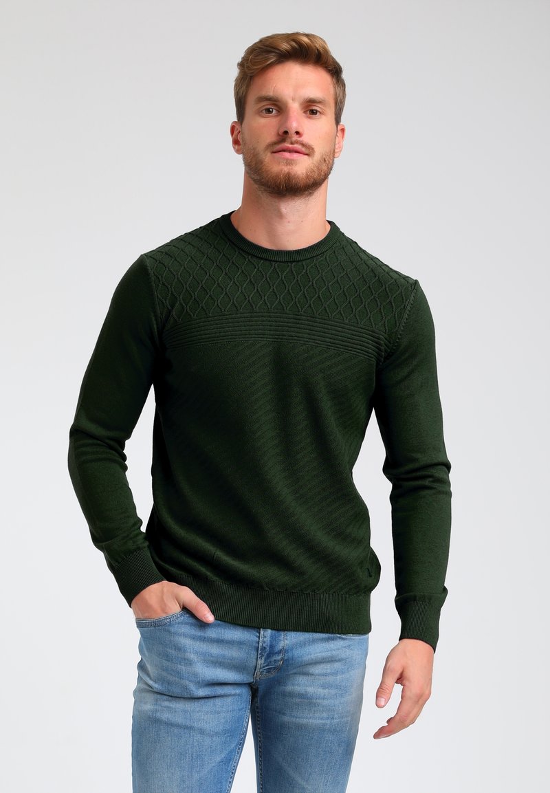 Donkergroen gebreid sweater met textuurpatronen op de bovenkant, geribbelde boorden en zoom, gecombineerd met lichtblauwe jeans.