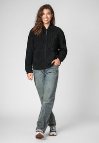 Schwarze Fleecejacke mit Reißverschluss vorne, einer Brusttasche und Seitentaschen; kombiniert mit hellblauen Jeans und Sneakers im Retro-Stil.
