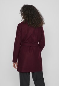 Burgundijas vilnas maisījuma mētelis ar jostu ap vidu un garām piedurknēm. Gluda tekstūra, taisns griezums un klasiskas formas apkakle aizmugurē.