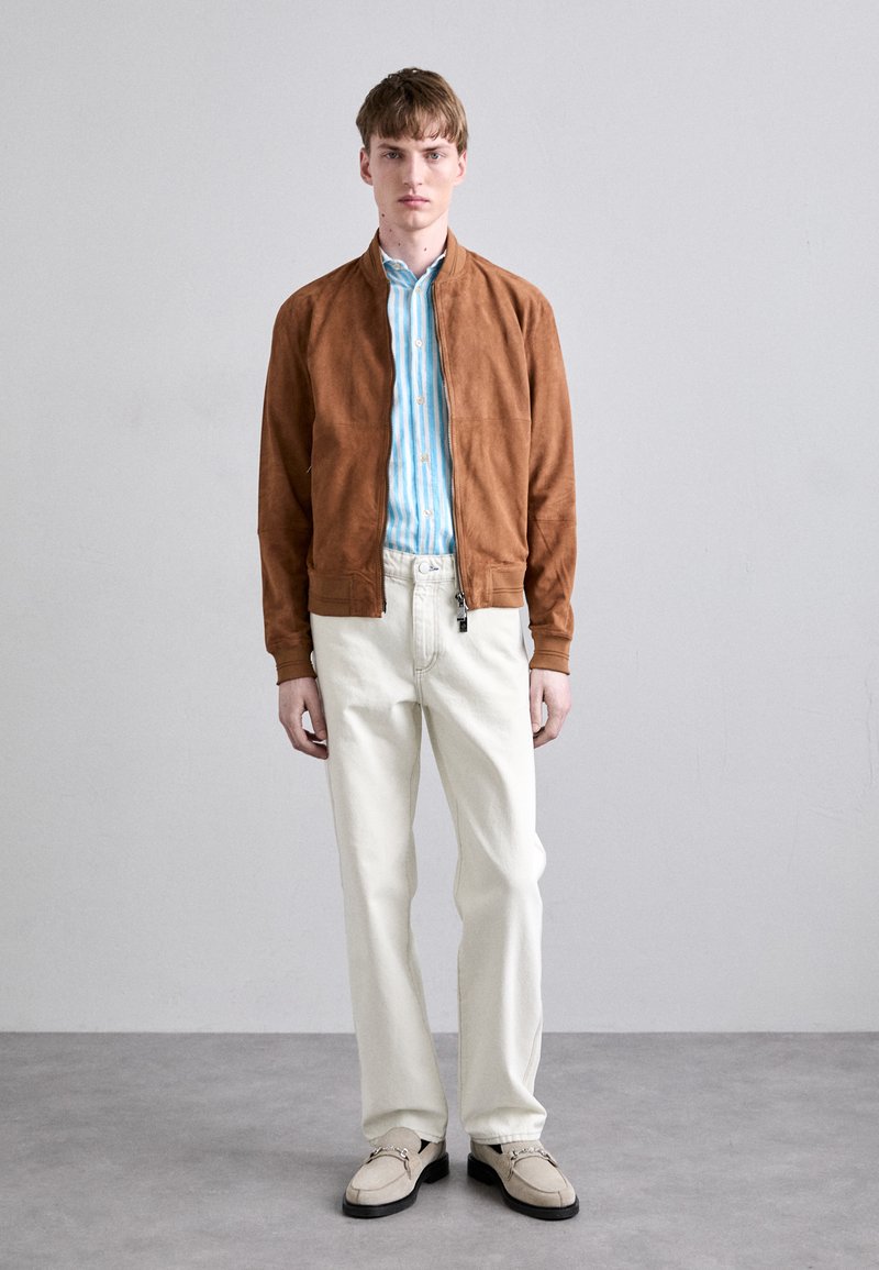 Chaqueta bomber de ante marrón sobre una camisa a rayas azul claro y blanca, combinada con pantalones crema y mocasines de color claro.