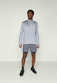 Mann står iført grå langermet treningsgenser, grå shorts med svarte kompresjonsshorts under, svarte sokker og svarte joggesko.