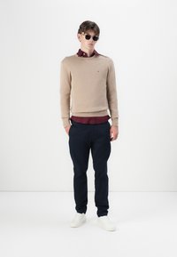 Beige kötött pulóver, bordó galléros inggel, bordázott mandzsettákkal, navy nadrággal és fehér sneakerrel párosítva. A modellen napszemüveg van.