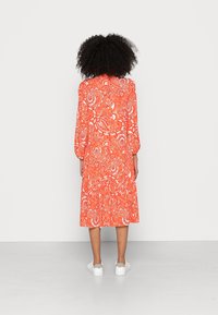 Rotes Kleid mit Paisleymuster, langen Ärmeln und gestuftem Saum, aus leichtem Stoff, kombiniert mit weißen Turnschuhen.