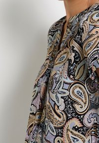 Kvinde iført en bluse med flerfarvet paisley-mønster i gule, blå, sorte og lavendelfarvede nuancer mod en ensfarvet baggrund.