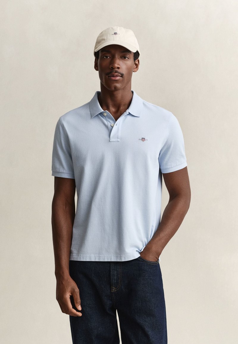 Homme portant un polo bleu clair, un jean foncé et une casquette de baseball claire, debout avec une main dans la poche devant un fond uni.