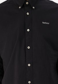 Fekete, gombos ing hegyes gallérral, fehér gombokkal és egy kis ezüst hímzett "Barbour" logóval a mellrészen.