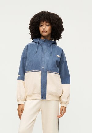 Kvinde med krøllet hår iført en blå og beige hættetrøje med snore og cremefarvede bukser, stående mod en ensfarvet baggrund.