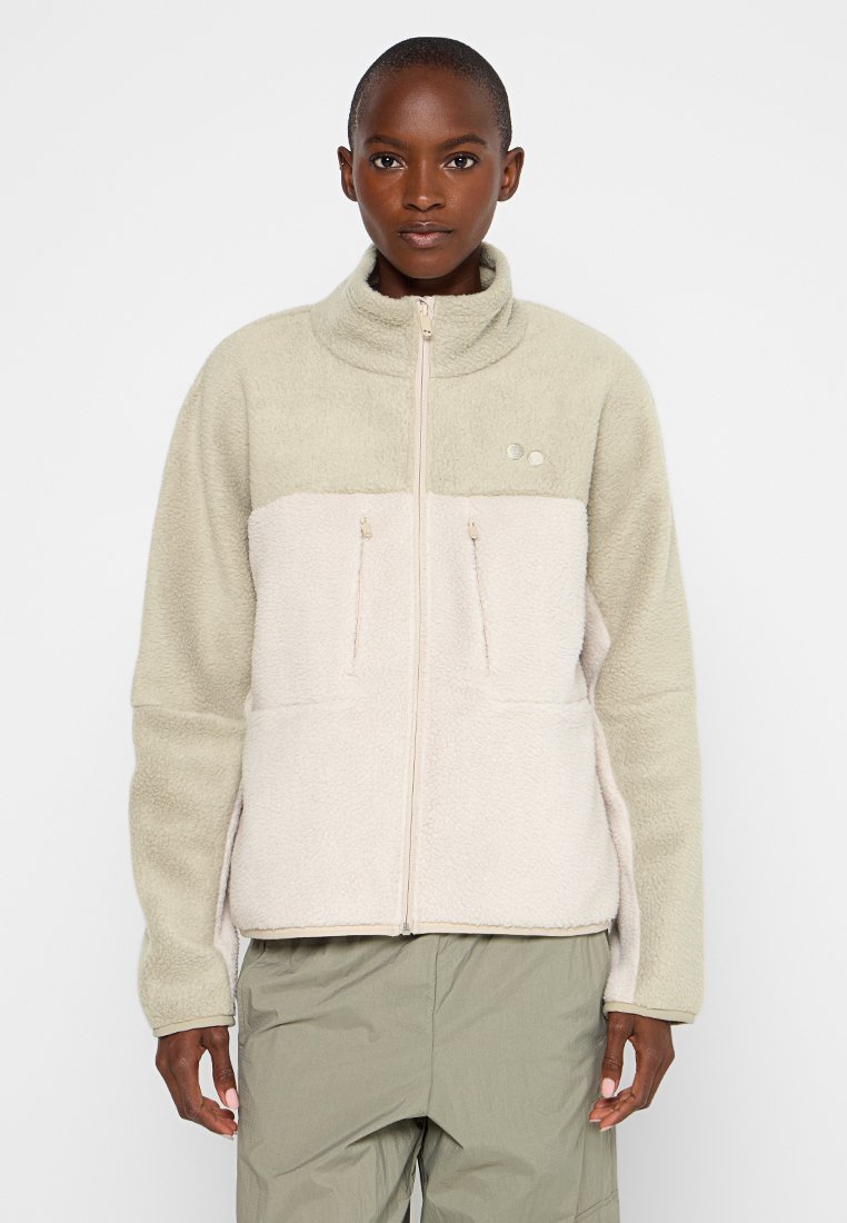 Fleecejacke mit hohem Kragen, mit einem beigen Oberteil und einem hellbeigen oberen Bereich. Mit Reißverschlusstaschen und dezentem Logodetail auf der Brust.