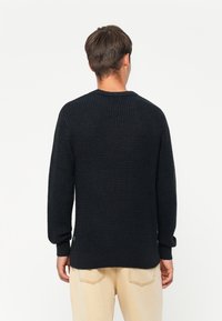 Uomo che indossa un maglione nero a trama e pantaloni beige chiaro, di spalle contro uno sfondo bianco uniforme.