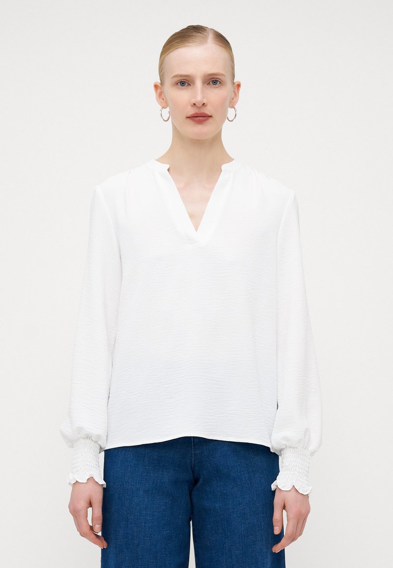 Weiße Bluse mit langen Ärmeln, V-Ausschnitt, strukturiertem Stoff und Rüschenmanschetten. Kombiniert mit dunkelblauen weit geschnittenen Jeans.