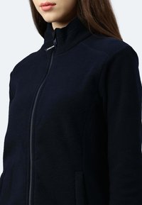 Giacca in pile blu navy con colletto alto, chiusura a zip e tasche laterali; texture morbida e design aderente evidenziato nella vista ravvicinata.