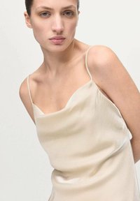 Camisola de seda color beige claro con tirantes finos; textura suave y sutil brillo. Escote redondo y ajuste relajado.