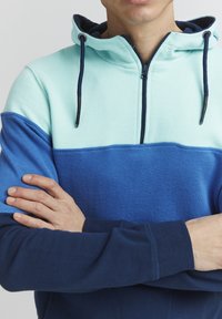 Mikina s kapucňou, ktorá obsahuje kombináciu svetlo zelenej, modrej a námorníckej modrej farby, má zips, textúrovanú tkaninu a detaily s nastaviteľným šnúrkami.