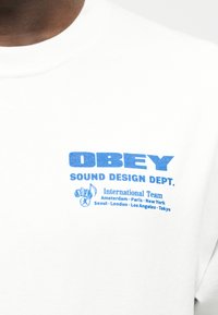 Άσπρο μπλουζάκι με μπλε κείμενο: «OBEY SOUND DESIGN DEPT. Διεθνής Ομάδα» και ονόματα πόλεων κάτω από ένα εικονίδιο μουσικής νότας και ακουστικών.